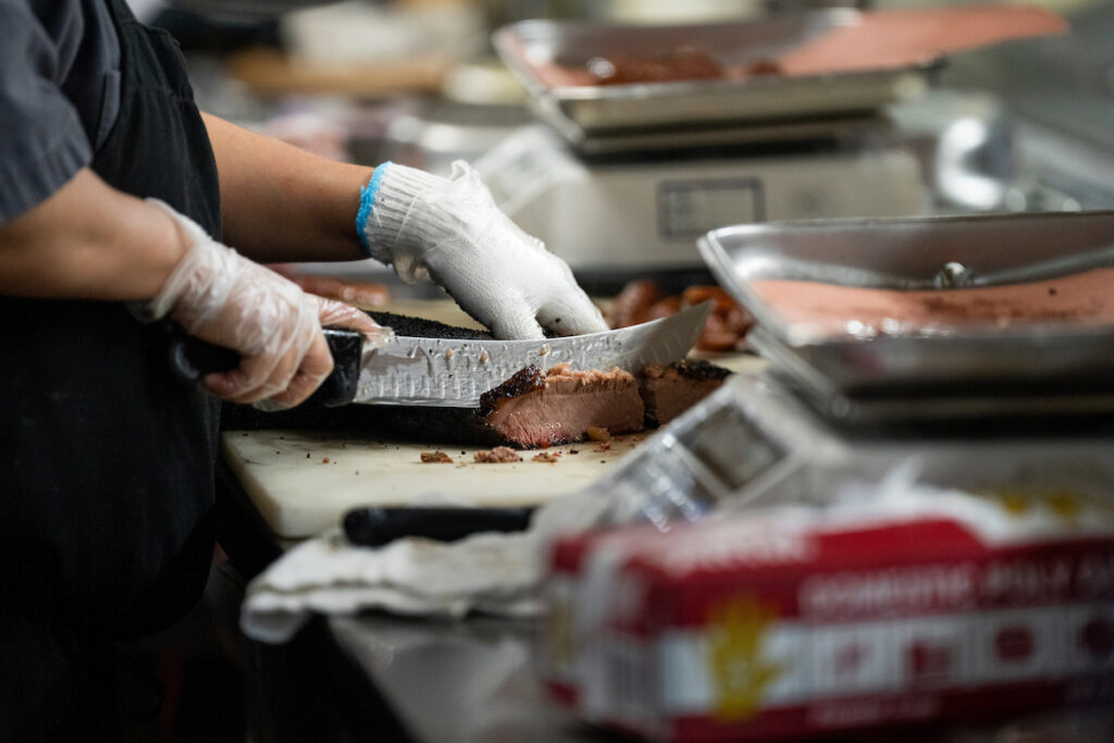 A pair of gloved hands slicing brisket on a cutting board next to two meat scales.