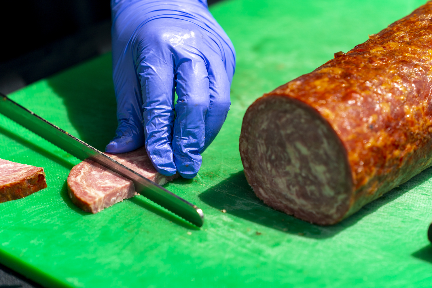 gloved hands slicing a dry sausage on a green cutting board