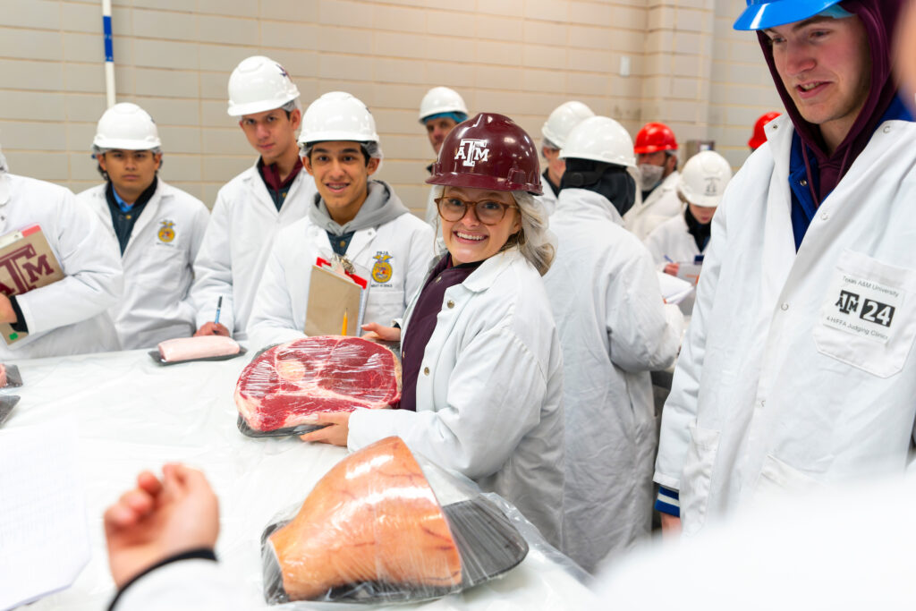 Several people wearing helmets and long white lab coats standing around table with two large cuts of meat.