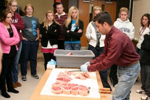 A group of people gathered around a table, watching a man who is handling various pieces of meat on the table.