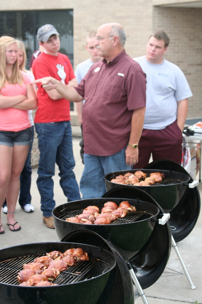 A professor standing with multiple grills cooking a lot of meat in each grill, while standing in front of a group of students. 