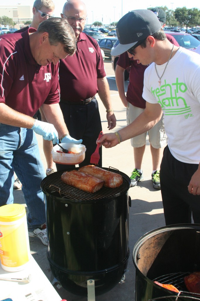 A group of men standing around a smoker, season the pieces of meat in the smoker. 