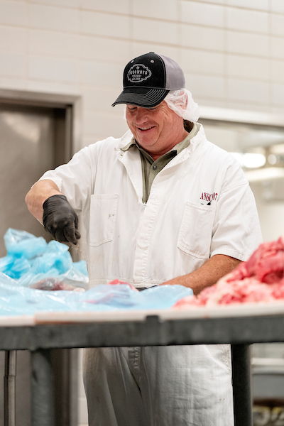 A man wearing gloves and kitchen safety gear stands in front of a table covered with blue bags and pieces of meat.