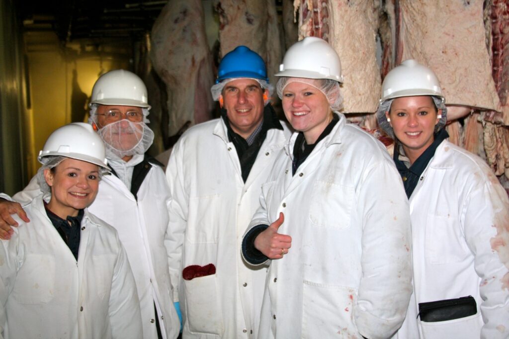 A group of people wearing hard hats and lab coats, with carcasses hanging in the background.