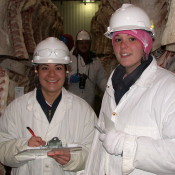 Two girls smelling at the camera wearing lab coats and hard hats. The one on the left is holding a clip board.