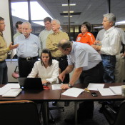 A meeting room, with multiple people discussing. There is one desk, with multiple pieces of paper, with a computer where two people are more closely discussing what on the computer.