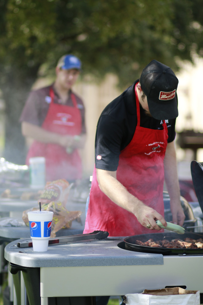 A guy wearing a red apron standing in front of a frill preparing some meat.