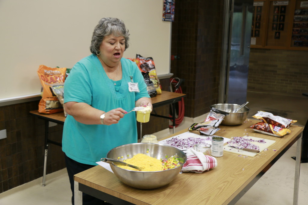 A lady standing next to a table preparing guacamole in a metal bowl. 