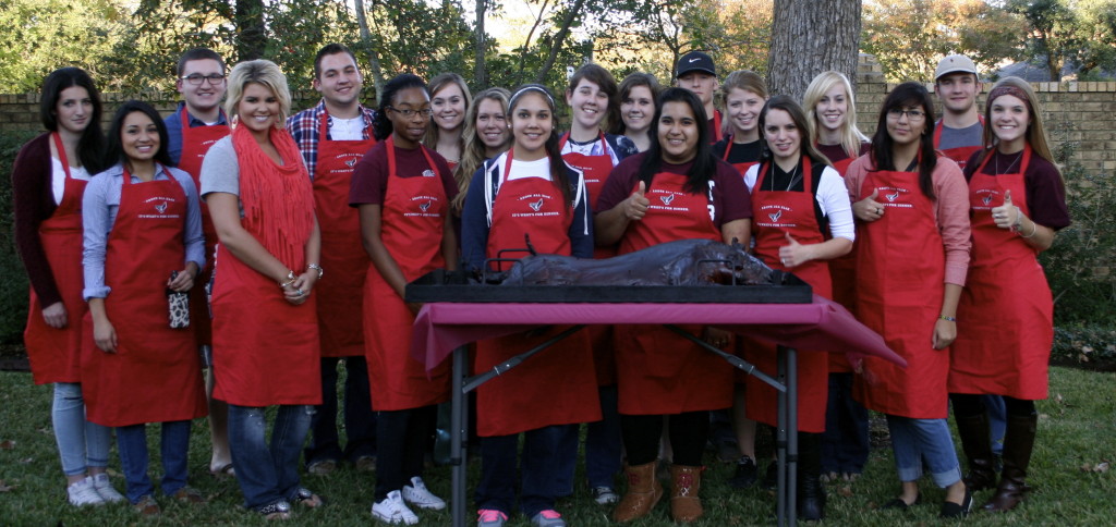 A group of students wearing red aprons standing in front of a table with a full pork on it. 