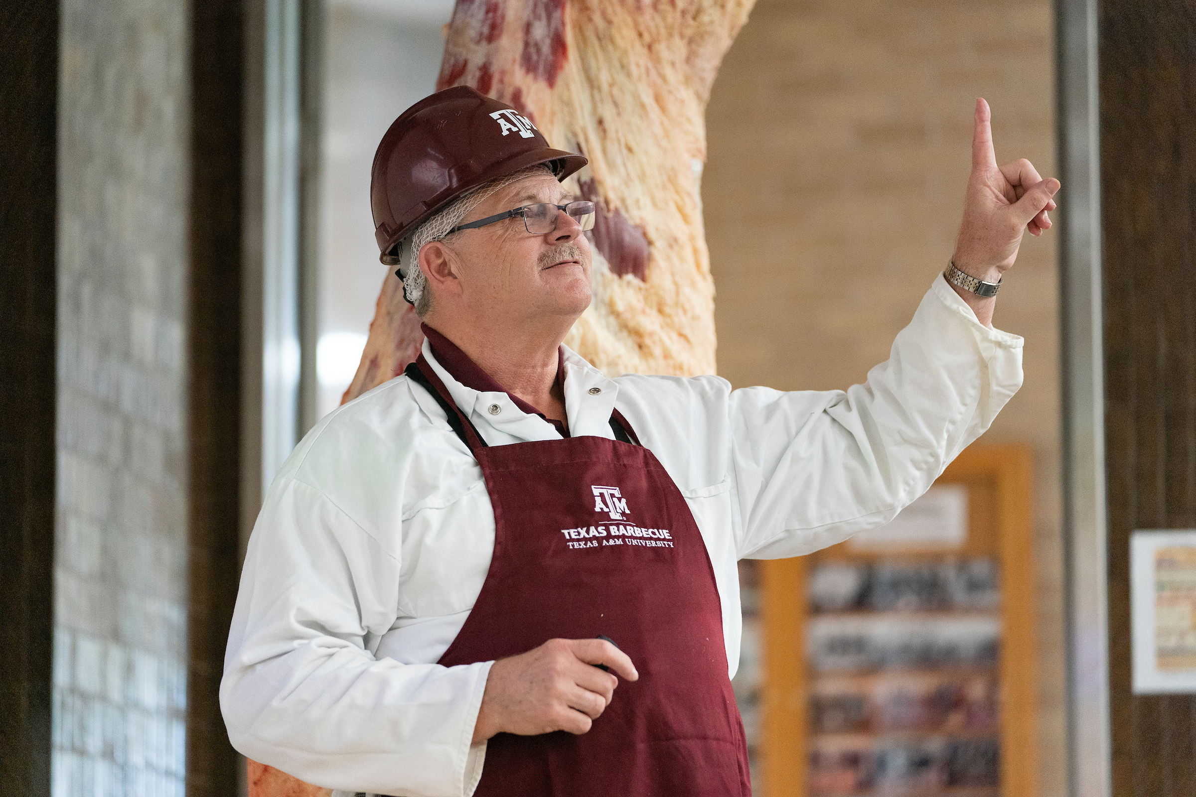 Man wearing helmet and apron speaks as he stands next to hanging meat carcass.