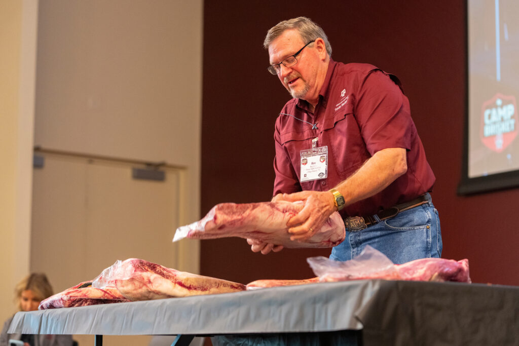 Man wearing name tag holding large cut of meat in front of classroom.