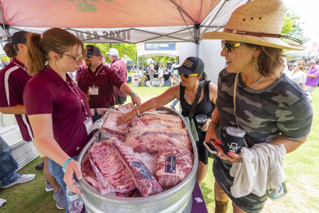 Two women examining a metal tub filled with ice and packaged racks of ribs, under a tent attended by three people in maroon polo shirts.