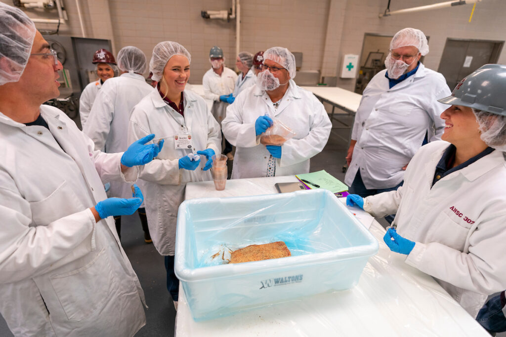 People wearing hair nets, blue gloves and coats, around a table with a plastic bin in the middle. 