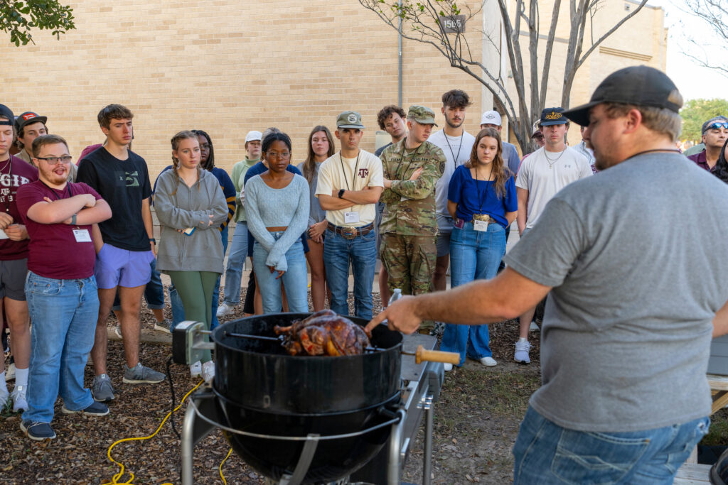  A professor is showing a piece of meat in a smoker to a group of student standing in front of him. 