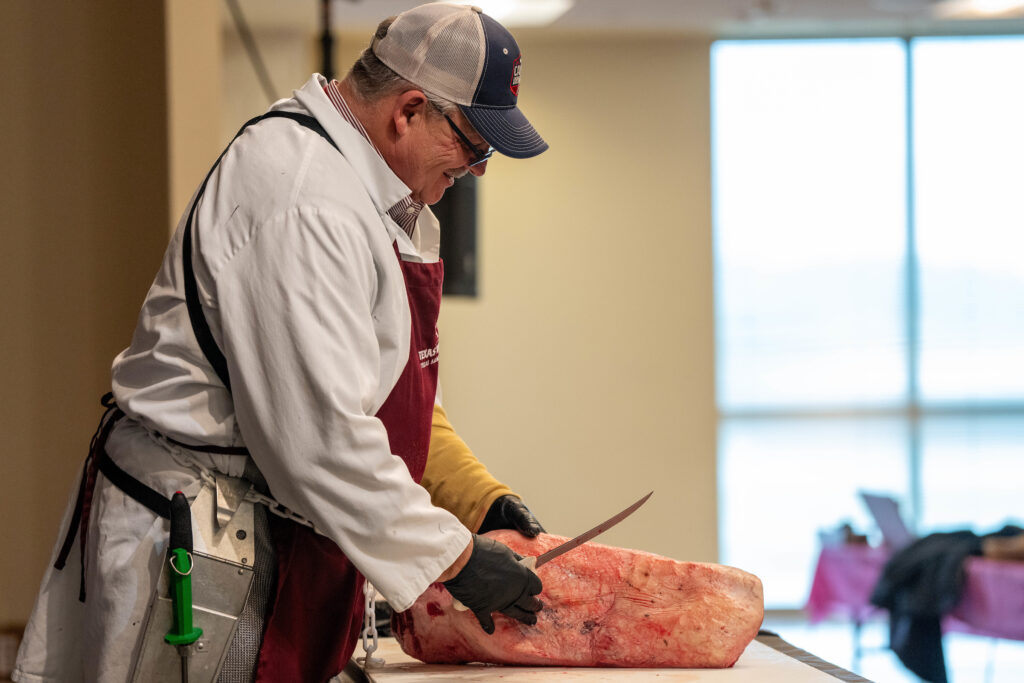 Man wearing cap and apron holding knife over large cut of meat in indoor setting.