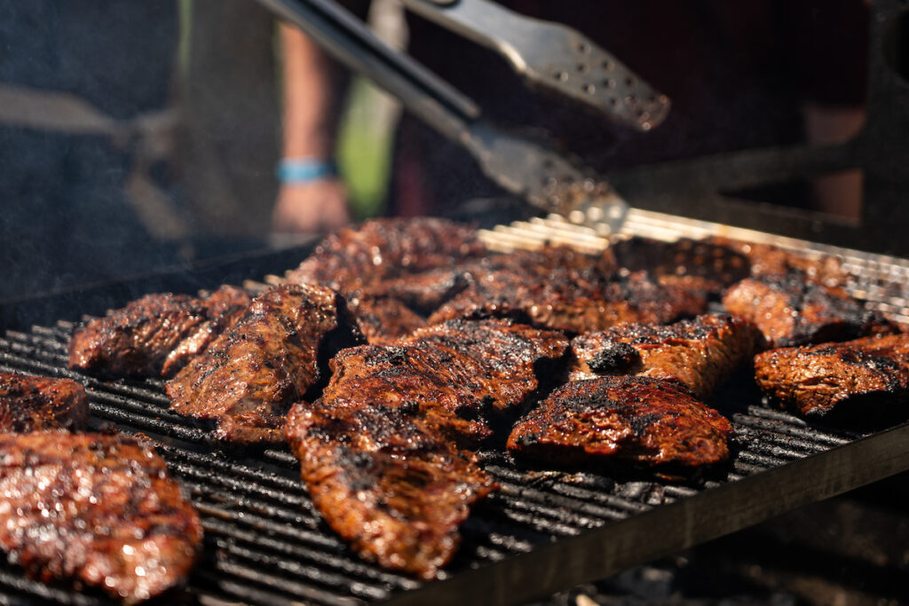 Several pieces of seasoned meat cooking on a large grill, with a pair of tongs reaching for one piece.